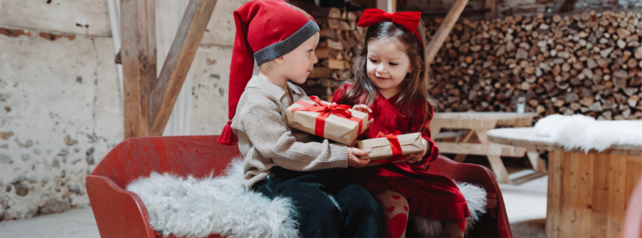 Two children in festive outfits sitting in a red sleigh holding a gift box.